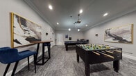 This single-floor amenity space is thoughtfully divided into distinct zones, each designed to serve a specific social or recreational purpose. The games room features patterned carpet flooring, crown molding, and recessed lighting complemented by a ceiling-mounted projector. It is equipped with a full-size pool table and a foosball table, along with high-top bar tables and seating along the perimeter. Framed artwork and wall-mounted sconces add a polished finish to the space.

Adjacent to the games room, the lounge area makes an immediate impression with its soaring ceilings, ornate fireplace with a decorative surround, and a bold navy accent wall hosting a wall-mounted flat-screen television. Built-in shelving flanks the fireplace, while a blue channel-tufted sofa, a geometric area rug, and marble-top side tables complete the seating arrangement. Large multi-pane windows flood the area with natural light.

The kitchenette features vaulted ceilings, white cabinetry, gray tile backsplash, stainless steel appliances including a refrigerator and microwave, and high-top tables with modern bar chairs. A smaller, quieter nook off the main areas provides a media retreat furnished with plush velvet lounge chairs, patterned carpet, and a wall-mounted screen — ideal for private viewing or relaxation.