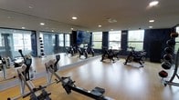 This well-appointed fitness studio is designed to support a focused and energetic workout environment. The space features a clean, open layout with light hardwood-style flooring that provides both durability and visual warmth. A row of stationary spinning bikes is positioned along the mirrored wall, allowing cyclists to monitor their form during sessions. A rowing machine occupies the foreground, offering a versatile full-body cardio option.

One wall is entirely covered by a large mirror, visually expanding the space and enhancing the sense of openness. A flat-screen television is mounted centrally on the mirror, providing entertainment or guided workout content during training sessions. Recessed lighting in the white ceiling supplements the generous natural light streaming in through a series of large windows along the perimeter, which frame views of surrounding greenery and contribute to a bright, airy atmosphere.

The dark-trimmed windows and walls create a sleek, contemporary contrast against the lighter flooring and ceiling. Additional equipment, including weighted medicine balls on a rack, can be spotted along the edges of the room, making this a well-rounded and functional fitness space.