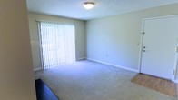 This single-floor space features a well-proportioned living area finished with wall-to-wall carpet in a neutral beige tone, contributing to a clean and comfortable atmosphere. The room is anchored by a large sliding glass door dressed with vertical blinds, which serves as the primary source of natural light and provides direct access to an outdoor area. The walls are painted in a soft, neutral tone that complements the light carpeting and promotes a bright, open feel. A flush-mount ceiling light fixture provides overhead illumination, and the textured ceiling adds a subtle architectural detail consistent with the overall understated design. White baseboards frame the perimeter of the room, offering a crisp contrast against the neutral walls and flooring. A transition to hard flooring is visible at one edge of the room, suggesting an adjoining space such as a kitchen or entryway. The layout is straightforward and functional, with ample open floor space suited for a variety of furniture arrangements. Overall, the space presents a simple, practical interior with natural light as a key feature.