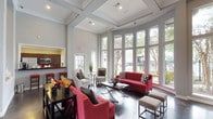 This lobby is a striking communal space defined by its dramatic architectural character and thoughtfully curated design. Soaring ceilings with coffered detailing and tall structural columns establish a grand sense of scale, while the polished concrete floors add an industrial-modern contrast to the otherwise classic aesthetic. Soft gray walls provide a neutral backdrop throughout.

Floor-to-ceiling windows line one side of the lobby, flooding the interior with abundant natural light and offering clear views of the surrounding landscape and outdoor greenery. The seating area is furnished with bold red sofas and accent chairs paired with gray throw pillows, arranged around low-profile coffee tables to create an inviting, social atmosphere. Neutral upholstered chairs with a side table form a quieter conversation nook near the windows.

A white brick fireplace serves as a prominent focal point along one wall, topped with a rustic wood mantle and a wall-mounted flat-screen television, framed by abstract wall art. French-paned interior doors connect to adjacent rooms, maintaining visual openness throughout. A kitchenette or café-style bar area is visible to one side, featuring dark cabinetry, appliances, and red bar stools, adding functional convenience to the space. Overall, the lobby presents a welcoming and polished environment suited for gathering and community interaction.
