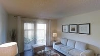 This single-floor space features an open-concept layout that connects the living area to the kitchen, creating a cohesive and functional flow. The living room is comfortably sized, finished with dark hardwood-style flooring and a textured ceiling. A large sliding glass door dominates one wall, fitted with sheer curtains and vertical blinds, filling the room with natural light and providing direct access to a private outdoor balcony. The living area accommodates a neutral-toned sofa, ottoman, and a glass coffee table, with framed artwork adding a refined touch to the walls. A media console in a metallic finish houses the television and decorative accents, anchoring the entertainment area. Adjacent to the living room, the kitchen is visible through an open passthrough, showcasing warm wood cabinetry, a full-size white refrigerator, and functional counter space. The overall ambiance is bright and understated, with neutral wall tones and natural light contributing to an airy, welcoming atmosphere throughout the space.