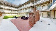 Located at 303 S Riverfront Dr in Jenks, Oklahoma, this multi-story residential community is designed around a central open-air courtyard that serves as the heart of the property. The courtyard features a large, lush artificial turf lawn bordered by a herringbone-patterned brick patio, creating a welcoming communal outdoor space. Adirondack chairs, outdoor dining furniture, and a fire pit area provide ample opportunities for relaxation and social gathering. A wood pergola structure adds a natural, rustic accent to one corner of the courtyard, enhancing its character and charm. The surrounding multi-story buildings showcase a clean, contemporary exterior with horizontal lap siding in neutral tones, complemented by dark-trimmed railings on the covered upper-level balconies. Ground-floor units open directly onto covered concrete walkways, with each entryway accented by a small patch of turf. The balconies on upper levels provide private outdoor space for residents while overlooking the vibrant courtyard below. Recessed lighting along the covered corridors ensures the walkways remain well-lit and functional. The cohesive architectural design, open courtyard layout, and thoughtfully arranged outdoor amenities combine to create a community-oriented living environment with a comfortable and inviting ambiance.