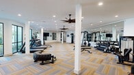 Located at 19101 Town Ridge Ln in Houston, TX, this generously sized fitness center spans a single floor and offers a well-appointed, professional-grade workout environment. The space is designed with an open layout that accommodates a diverse array of equipment zones, allowing members to seamlessly transition between cardio, strength training, and functional fitness areas.

The flooring features a distinctive herringbone-patterned carpet in warm gold and cool gray tones, lending both visual interest and practical durability underfoot. Large black-framed windows line portions of the perimeter, inviting abundant natural light and offering views of the surrounding greenery. Multiple ceiling fans are suspended from the bright white ceiling alongside recessed lighting, ensuring effective air circulation and consistent illumination throughout the space.

Floor-to-ceiling mirrors are strategically positioned along several walls, visually expanding the room and providing members with proper form-checking capability. Equipment includes treadmills, ellipticals, stationary and spin bikes, rowing machines, a cable machine, Smith machine, multi-use weight bench, a full dumbbell rack, kettlebells, medicine balls, stability balls, and a large training tire for functional workouts. A wall-mounted flat-screen TV adds an entertainment element to the space. The clean, white interior palette creates a bright, energizing atmosphere suited for a high-quality fitness experience.