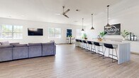 Located at 316 Columbia Dr in Carrollton, GA, this single-floor game room — branded as The Foundry — offers a generously sized, open-concept layout designed for socializing and recreation. The space is finished with light wood-look flooring throughout, crisp white walls, and recessed lighting complemented by stylish pendant fixtures and a ceiling fan, creating a clean, modern aesthetic.

The main area is thoughtfully divided into distinct activity zones. A large, plush sectional sofa anchors the lounge area, paired with a wall-mounted flat-screen TV, making it ideal for group gatherings. Adjacent to the lounge, a full-length bar counter lined with sleek black barstools and illuminated by industrial-style pendant lights serves as a central hub, highlighted by The Foundry signage above. A regulation-size pool table with a blue felt surface occupies its own dedicated zone, complete with cue racks and overhead pendant lighting.

Numerous windows along multiple walls flood the interior with natural light, offering views of the surrounding outdoor greenery and providing an airy, open feel. An additional flexible area near the entry features clean white walls and direct access to the exterior. The overall ambiance is contemporary and inviting, well-suited for both casual recreation and community events.