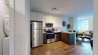 This well-appointed one-bedroom, one-bathroom unit offers a thoughtfully designed layout across a single floor, making efficient use of every square foot. The kitchen is a standout feature, showcasing high-gloss white upper cabinetry that extends to the ceiling for maximum storage, contrasted by dark wood lower cabinets. A full suite of stainless steel appliances — including a refrigerator, range, microwave, and dishwasher — pairs with a light quartz countertop and a peninsula that doubles as a breakfast bar. Rich hardwood-style flooring runs throughout the main living areas, adding warmth and continuity. The living room is generously lit by floor-to-ceiling windows that frame impressive city skyline views, creating a bright and airy atmosphere. The bedroom offers a comfortable retreat, complete with a dedicated closet, natural light from a window with urban views, and warm ambient lighting. The bathroom is finished with large-format tile flooring, a dark wood vanity with a white countertop, and a shower-tub combination. A stacked washer and dryer unit is also conveniently included within the space, adding a practical everyday amenity to this modern urban residence.