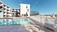 Located in the heart of Hollywood at 7059 Willoughby Ave, this rooftop amenity deck offers a vibrant outdoor retreat with a distinctly urban Los Angeles character. The centerpiece of the space is a generously sized lap pool with crystal-clear blue water, framed by clean, large-format tiled decking and lined with lounge chairs perfect for soaking up the Southern California sun. String lights are strung overhead, lending a festive ambiance that transitions effortlessly from daytime relaxation to evening gatherings. An outdoor kitchen station anchors one end of the deck, featuring a stainless steel grill and concrete countertops accented by colorful mosaic tile work in soft yellow, blue, and white tones. Glass panel railings around the perimeter preserve unobstructed panoramic views of the surrounding Los Angeles skyline, including neighboring mid-rise buildings and distant mountain silhouettes. A separate courtyard-level terrace is visible below, equipped with outdoor seating and lush plantings, providing an additional communal gathering area. The modern architecture of the building — characterized by crisp white facades, open balconies, and expansive glazing — complements the bright, open character of the rooftop deck, creating a polished and contemporary outdoor living environment.