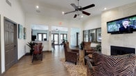This single-story space presents an open, interconnected floor plan that flows seamlessly between a welcoming lounge area, a dining or activity room, and what appears to be a front reception or service counter. The main lounge is anchored by a textured area rug and furnished with striking zebra-print upholstered armchairs and leather seating, lending the space a bold, eclectic character. A ceiling fan with integrated lighting is centrally mounted overhead, complementing the recessed lighting throughout and contributing to a well-lit, comfortable atmosphere.

The flooring is consistent hardwood-style planking that unifies the open zones visually. Arched and rectangular doorways create natural transitions between areas without the use of doors, maintaining an airy, connected feel. The adjacent room, visible through a wide opening, features bar-height seating and large glass doors that invite natural light and suggest views to an outdoor area.

To the right, a stone or tile-clad feature wall adds architectural texture and anchors a large framed mirror, enhancing the sense of depth. Tropical plants placed throughout introduce a fresh, lively element. The overall ambiance is casual yet characterful, suited for social gatherings or community use.