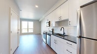 Located at 1636–1648 NW 38th St in Kansas City, Missouri, this modern single-floor unit offers a clean, contemporary design throughout. The open-concept living and kitchen area is anchored by large windows that flood the space with natural light and frame views of the surrounding Kansas City cityscape. Wood-look flooring runs consistently throughout, lending warmth and visual cohesion to the layout.

The kitchen is well-appointed with crisp white cabinetry, matte black hardware, dark granite countertops, a white subway tile backsplash, and a full suite of stainless steel appliances including a range, microwave, dishwasher, and refrigerator. A deep undermount sink with a black matte faucet completes the functional yet stylish workspace.

The living area is generously sized, with wide windows offering elevated views of the neighborhood. The bedroom continues the bright, airy aesthetic with its own window framing outdoor scenery and ample floor space. The bathroom features white vanity cabinetry with dark countertops, a large mirror, and a tiled shower enclosure. An in-unit stacked washer and dryer adds everyday convenience. The overall ambiance is modern, light-filled, and move-in ready.