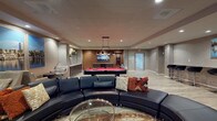 Located at 270 Pacific Ave in Long Beach, CA, this single-floor amenity lounge is thoughtfully designed to serve as a versatile social and recreational space. The interior features light wood-look flooring throughout, a drop tile ceiling with recessed lighting, and a neutral color palette accented by warm earth tones on select walls, creating a welcoming and modern atmosphere.

The central lounge area is anchored by a large curved black leather sectional sofa paired with a round glass coffee table, positioned atop a patterned area rug. Opposite the seating, a full-size red felt pool table commands attention beneath a set of industrial-style pendant lights, with a wall-mounted flat-screen TV positioned behind it. A built-in shelving unit nearby adds additional display and storage.

Along one wall, a countertop bar ledge lined with sleek black swivel bar stools provides additional seating, accompanied by a nautical-themed canvas print above. A wet bar kitchenette features dark wood cabinetry, a granite countertop, a sink, a compact refrigerator, and floating shelves with decorative accents. A secondary seating nook offers gray upholstered chairs arranged around a triptych cityscape artwork depicting Long Beach's waterfront skyline, adding a locally inspired finishing touch.