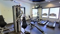 This dedicated fitness center is designed to support a well-rounded workout routine in a comfortable, purpose-built environment. The room's most distinctive architectural feature is its tray ceiling, finished with a deep navy blue inset that adds a striking visual accent overhead. A ceiling fan is centrally mounted within the tray, promoting air circulation throughout the space, while a wall-mounted flat-screen television provides entertainment during workouts.

The fitness center is outfitted with a thoughtfully curated selection of exercise equipment. A Precor multi-station cable and weight machine anchors one side of the room, offering a wide range of strength training options. The cardio section features an elliptical trainer and at least two treadmills, all positioned to take advantage of the natural light filtering in through multiple windows fitted with horizontal blinds. The windows provide views to the outdoors, contributing to an open, energizing atmosphere.

Durable rubber flooring covers the entire space, offering both safety and function underfoot. The neutral gray walls paired with the bold ceiling detail create a clean, modern aesthetic. Overall, the fitness center is bright, practical, and well-maintained, making it an inviting space for daily exercise.
