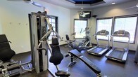 This dedicated fitness center is designed to support a well-rounded workout routine in a comfortable, purpose-built environment. The room's most distinctive architectural feature is its tray ceiling, finished with a deep navy blue inset that adds a striking visual accent overhead. A ceiling fan is centrally mounted within the tray, promoting air circulation throughout the space, while a wall-mounted flat-screen television provides entertainment during workouts.

The fitness center is outfitted with a thoughtfully curated selection of exercise equipment. A Precor multi-station cable and weight machine anchors one side of the room, offering a wide range of strength training options. The cardio section features an elliptical trainer and at least two treadmills, all positioned to take advantage of the natural light filtering in through multiple windows fitted with horizontal blinds. The windows provide views to the outdoors, contributing to an open, energizing atmosphere.

Durable rubber flooring covers the entire space, offering both safety and function underfoot. The neutral gray walls paired with the bold ceiling detail create a clean, modern aesthetic. Overall, the fitness center is bright, practical, and well-maintained, making it an inviting space for daily exercise.