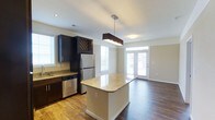 This single-floor residence showcases a thoughtfully designed open-concept layout that seamlessly connects the kitchen and living areas. The kitchen is outfitted with dark espresso cabinetry, stainless steel appliances including a refrigerator and dishwasher, and granite countertops that extend to a central island. The island, illuminated by a contemporary rectangular pendant light, serves as both a functional workspace and a casual gathering point.

Dark hardwood-style flooring runs throughout, providing a warm contrast to the light-toned walls and granite surfaces. A decorative lattice-panel cabinet above the refrigerator adds a distinctive architectural detail to the kitchen. The living area beyond benefits from abundant natural light, courtesy of multiple windows and a set of French doors that open to the exterior, creating an airy, welcoming atmosphere. Crown molding frames the living space, lending a refined finish. The overall design balances practical functionality with modern aesthetics, resulting in a cohesive and inviting environment.