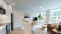 This well-appointed single-floor residence showcases a thoughtfully designed open-concept layout that seamlessly connects the kitchen, dining, and living areas. The kitchen is outfitted with sleek white cabinetry, warm wood open shelving, white quartz countertops, a tile backsplash, and a full suite of stainless steel appliances. A white waterfall-edge island with bar seating serves as both a functional prep space and casual dining area. The living room is bathed in natural light from floor-to-ceiling windows that frame urban views, and features light hardwood flooring that runs throughout the home. A striking accent wall with a flowing, marbled wallpaper mural adds a distinctive design element to the living space. The bedroom is a serene retreat, offering large windows, a sliding closet, and continued hardwood flooring. The bathroom is clean and modern, featuring a single-sink vanity with a lit mirror, glass-enclosed walk-in shower with tile surround, and wood-look flooring. Neutral tones, recessed lighting, and consistent contemporary finishes create a cohesive, bright, and comfortable atmosphere throughout the entire space.