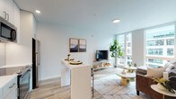 This well-appointed single-floor residence showcases a thoughtfully designed open-concept layout that seamlessly connects the kitchen, dining, and living areas. The kitchen is outfitted with sleek white cabinetry, warm wood open shelving, white quartz countertops, a tile backsplash, and a full suite of stainless steel appliances. A white waterfall-edge island with bar seating serves as both a functional prep space and casual dining area. The living room is bathed in natural light from floor-to-ceiling windows that frame urban views, and features light hardwood flooring that runs throughout the home. A striking accent wall with a flowing, marbled wallpaper mural adds a distinctive design element to the living space. The bedroom is a serene retreat, offering large windows, a sliding closet, and continued hardwood flooring. The bathroom is clean and modern, featuring a single-sink vanity with a lit mirror, glass-enclosed walk-in shower with tile surround, and wood-look flooring. Neutral tones, recessed lighting, and consistent contemporary finishes create a cohesive, bright, and comfortable atmosphere throughout the entire space.