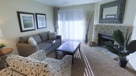 This single-story space centers around a well-appointed living room that balances comfort and style. A large sliding glass door fitted with vertical blinds allows abundant natural light to filter into the room, casting bright streaks across the carpeted floor. The living area is anchored by a gray sofa and coordinating patterned accent chairs, arranged around a dark leather-topped coffee table that serves as a functional centerpiece. A standout feature of the room is the stone fireplace situated in the corner, adding a rustic, textured contrast to the otherwise neutral, modern décor. The warm-toned stone hearth extends to the floor, creating a cozy focal point ideal for gathering. Decorative framed artwork lines one wall, contributing a gallery-like feel, while an ornate framed mirror above the fireplace mantle enhances the sense of depth and openness. Tall decorative branches in a floor vase add a natural, organic element. The neutral color palette—encompassing soft grays, beige carpeting, and warm white walls—creates a calm, welcoming atmosphere throughout the space.