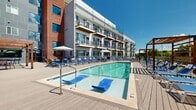 Located at 947 Myers St in Richmond, Virginia, this outdoor amenity deck offers a well-appointed resort-style retreat designed for relaxation and recreation. The centerpiece of the space is a generously sized swimming pool with crystal-clear water, complemented by a shallow wading area outfitted with floating lounge chairs — ideal for unwinding in the sun. Surrounding the pool, a broad composite wood deck provides ample room for lounging, with rows of blue chaise lounge chairs and market umbrella stations arranged throughout. Warm-toned wooden pergolas add architectural character to the deck, offering shaded gathering spots with seating for dining and socializing. String lights are strung between structures, lending an inviting ambiance for evening use. The deck is enclosed by a sleek railing system, with mature greenery and open Richmond skyline views visible beyond the perimeter. The adjacent multi-story building features a modern facade combining brick, metal panel, and large-format windows with private balconies that overlook the pool area. The overall design blends contemporary architecture with approachable outdoor living, creating a vibrant communal space that captures abundant natural light and open skies throughout the day.