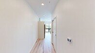 This well-designed 1-bedroom, 1-bathroom unit offers a thoughtful layout across a single floor, making efficient use of its compact footprint. The combined kitchen and living area serves as the heart of the space, featuring light hardwood flooring throughout, recessed lighting, and a striking wall of large, black-framed windows that flood the room with natural light and frame views of the surrounding greenery. The kitchen is outfitted with sleek white cabinetry, dark countertops, a full suite of stainless steel appliances including a refrigerator, range, microwave, and dishwasher, and a central island with a marble-top surface ideal for dining or meal prep. A clean hallway with recessed ceiling lights connects the main living area to the private bedroom and bathroom. The bedroom is a calm, neutral space finished with soft carpeting and a window for additional natural light. The bathroom features a walk-in shower, a white vanity with an integrated sink, a large mirror, and brushed nickel fixtures, maintaining the unit's clean, contemporary aesthetic. A dedicated closet provides practical storage. Throughout, the crisp white walls, consistent modern finishes, and cohesive design create a bright and functional living environment.