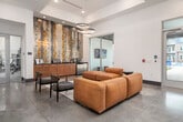 This contemporary single-floor amenity space is designed with a clean, modern aesthetic that immediately impresses upon entry. The vestibule entrance features dark-framed glass doors with transom windows that flood the space with natural light, offering views of the surrounding greenery outside.

The main lounge area is anchored by a dramatic accent wall adorned with a textured, gold and silver tonal art installation, set above a warm wood credenza that serves as a beverage or service station. This focal point is framed by a tray ceiling detail and illuminated by a pair of elegant circular pendant light fixtures in brushed gold, adding a sophisticated layer of ambient lighting.

Seating throughout the lounge consists of rich caramel-toned leather sectional sofas arranged atop a geometric patterned area rug, complemented by dark upholstered accent chairs with sleek metal frames. Polished concrete flooring extends throughout, lending an industrial-chic character to the overall design.

The space benefits from abundant natural light through large glass doors and windows, with direct access to an outdoor patio area. A fitness center is also accessible from within the lounge, visible through glass partitions. The high ceilings, open layout, and thoughtful furniture arrangement create an inviting, functional environment suited for both relaxation and community gatherings.