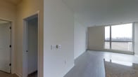 This single-floor space offers a straightforward, functional layout designed with clean lines and a neutral aesthetic. The interior is finished with smooth, white walls and crisp white baseboards that lend a bright, modern feel throughout. Durable hard-surface flooring in a cool gray-toned wood-look finish runs consistently across the main areas, providing both visual continuity and easy maintenance. A hallway connects the main living area to additional rooms, hinting at a well-organized floor plan. The living area is highlighted by a large window that draws in generous natural light, creating an open and airy atmosphere. The window's expansive scale suggests elevated views of the surrounding environment. Walls are equipped with modern switch plates and outlets, reflecting a thoughtfully prepared, move-in-ready condition. The space is presented in an unfurnished state, allowing viewers to clearly appreciate the clean architecture, proportions, and flow between areas. Overall, the space communicates a minimalist, contemporary character well-suited for a variety of uses and personalization.
