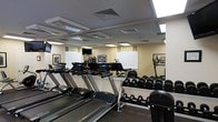 This single-floor fitness center is thoughtfully designed to accommodate a wide range of exercise needs. The space is anchored by a row of modern treadmills, each equipped with individual digital displays for tracking performance metrics. A dedicated free weights area lines the walls, featuring an organized dumbbell rack with a full range of weights, making strength training accessible for all fitness levels. Mirrored walls run along the perimeter, creating a sense of expanded space while allowing users to monitor their form during workouts. The ceiling features recessed lighting panels that provide bright, even illumination throughout the room. Mounted televisions are positioned for visibility from the cardio equipment, enhancing the workout experience. Natural light filters in through several windows fitted with blinds, complementing the artificial lighting. Framed black-and-white artwork decorates the walls, adding a polished aesthetic touch to the utilitarian space. The flooring is finished with a durable, dark rubberized surface suited for high-traffic gym use. Ventilation is provided through ceiling-mounted HVAC units, ensuring a comfortable environment during intense workouts. Overall, the space strikes a balance between functionality and a clean, professional atmosphere.