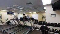 This single-floor fitness center is thoughtfully designed to accommodate a wide range of exercise needs. The space is anchored by a row of modern treadmills, each equipped with individual digital displays for tracking performance metrics. A dedicated free weights area lines the walls, featuring an organized dumbbell rack with a full range of weights, making strength training accessible for all fitness levels. Mirrored walls run along the perimeter, creating a sense of expanded space while allowing users to monitor their form during workouts. The ceiling features recessed lighting panels that provide bright, even illumination throughout the room. Mounted televisions are positioned for visibility from the cardio equipment, enhancing the workout experience. Natural light filters in through several windows fitted with blinds, complementing the artificial lighting. Framed black-and-white artwork decorates the walls, adding a polished aesthetic touch to the utilitarian space. The flooring is finished with a durable, dark rubberized surface suited for high-traffic gym use. Ventilation is provided through ceiling-mounted HVAC units, ensuring a comfortable environment during intense workouts. Overall, the space strikes a balance between functionality and a clean, professional atmosphere.