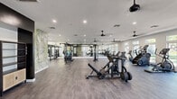 This expansive fitness center spans a generously sized single floor, thoughtfully designed to accommodate a comprehensive range of workout activities. The space is divided into distinct functional zones, each catering to different training needs. The strength training area features multiple cable machines, free-weight stations, barbells, and adjustable benches, all reflected in full-length mirror walls that enhance the sense of openness. A dedicated free weights section includes an organized dumbbell rack and functional training rig with TRX suspension equipment. The cardio zone, positioned along a wall of large windows, houses stationary bikes, ellipticals, a stair climber, and rowing machines, benefiting from ample natural light and outdoor views of surrounding greenery. Decorative tropical botanical murals serve as bold accent walls throughout, adding a vibrant, resort-inspired aesthetic that contrasts with the clean white walls and ceilings. Durable flooring—varying between wood-look planks and rubberized surfaces—is used strategically across zones. Multiple ceiling fans with recessed lighting maintain air circulation and illuminate the space evenly. A floor-to-ceiling glass entry door provides a welcoming, light-filled entrance.