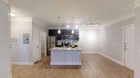 This single-floor space showcases a thoughtfully designed open-concept layout, centered around a well-appointed kitchen that serves as the heart of the living area. The kitchen features a large central island with a granite countertop, offering both a functional workspace and a casual gathering point. Dark espresso-toned cabinetry provides a striking contrast against the light-toned walls, while stainless steel appliances — including a refrigerator, microwave, and range — lend a modern, polished aesthetic.

Pendant lighting hangs above the island, complemented by a track lighting system that illuminates the space efficiently. Crown molding lines the ceiling throughout, adding a refined architectural detail that elevates the overall design. The light hardwood flooring flows seamlessly across the open area, contributing to a bright and airy atmosphere.

A paneled door visible to the side suggests additional rooms beyond the main living and kitchen area. The neutral grey-toned walls and white trim create a clean, cohesive palette that allows the darker cabinetry and granite surfaces to stand out. Overall, the space balances contemporary finishes with classic detailing for a welcoming and functional environment.