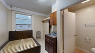 Located at 710 S Broadway in the heart of Downtown Los Angeles, this single-floor studio unit offers an efficient and functional layout designed to maximize every inch of available space. The main living area doubles as a sleeping and living space, featuring light wood-style laminate flooring that runs throughout, neutral beige walls, and a textured ceiling. A window positioned along one wall allows natural light to filter in, offering a view of the surrounding urban brick exterior. A wall-mounted heater unit provides in-unit climate control. The compact kitchen area is integrated into the main living space and includes a two-burner cooktop with a built-in sink, dark wood cabinetry below for storage, and upper wooden cabinets for additional pantry space. A solid wood entry door with a gold-tone handle opens into a small entryway that leads directly into the main room. The private bathroom is situated just off the main living area and is equipped with a pedestal-style sink, a wall-mounted mirror with an overhead medicine cabinet, a standard toilet, and a towel bar. The bathroom continues the light flooring aesthetic, maintaining a cohesive look throughout the unit. This studio is a practical urban living option in one of Los Angeles' most centrally located neighborhoods.