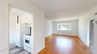 This single-floor residence offers a clean, functional layout with a consistent design language carried throughout. Warm-toned hardwood floors run through the living areas, bedrooms, and connecting spaces, lending continuity and character to the interior. White walls and crisp trim provide a bright, neutral backdrop across every room.

The living area is generously proportioned and receives ample natural light through large sliding windows. The kitchen features a gas range, white cabinetry, and an open pass-through that connects it to the main living space. A separate utility or service area includes a tiled countertop with white cabinetry above and below, accented by a decorative floral tile border, with light wood-look flooring and exterior door access.

The bedrooms are well-sized and sunlit, with large windows in each — one corner bedroom benefits from dual-aspect windows framing views of mature palm and tropical trees, flooding the space with natural light and greenery. Built-in closets with upper cabinetry and shelving provide practical storage solutions in the bedrooms, while ceiling fans add comfort.

The bathroom retains a vintage charm with warm terracotta-toned square tiles lining the tub surround, finished with a green accent border. A wall-mounted heating unit in the hallway area and panel doors throughout complete the space's cohesive, understated aesthetic.