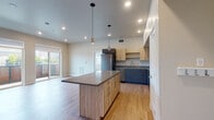 This single-floor residence showcases a contemporary design with light hardwood-style flooring throughout and a neutral color palette that creates a clean, cohesive aesthetic. The open-concept kitchen and living area serves as the heart of the unit, anchored by a sizeable center island with a wood-tone finish and dark countertop. The kitchen is well-appointed with two-tone cabinetry—pairing navy lower cabinets with natural wood upper cabinets and open shelving—complemented by a full suite of stainless steel appliances including a refrigerator, range, microwave, and dishwasher. Pendant lighting and recessed fixtures illuminate the space effectively. Sliding glass doors in the living area open to a balcony, bringing in generous natural light and urban views. The bedrooms are comfortably proportioned; one features dual windows that flood the room with sunlight and city views, while another includes a walk-in closet with built-in shelving. Two full bathrooms each feature a tub-shower combination with white tile surrounds, modern gray vanities, and wood-look flooring, maintaining the unit's consistent modern style throughout.