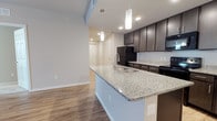 This single-floor residence features three bedrooms and two bathrooms across a thoughtfully arranged floor plan. The kitchen serves as a central focal point, showcasing a large granite-topped island with an integrated stainless steel sink and seating potential. Sleek dark espresso cabinetry lines the walls, complemented by matching dark appliances and consistent granite countertops that extend throughout the cooking area. Pendant lighting above the island adds a modern touch, while recessed ceiling lights provide even illumination across the space.

The kitchen opens directly into the living area, creating a connected, open-concept flow ideal for everyday living and entertaining. Durable wood-look flooring runs throughout the main living spaces, contributing to a cohesive and contemporary aesthetic. The neutral gray and white color palette throughout enhances the sense of openness and light. A hallway visible from the kitchen leads toward the bedrooms, suggesting a practical and logical layout that separates sleeping areas from common spaces. Overall, the unit presents a clean, modern design with quality finishes and a functional arrangement suited for comfortable daily living.