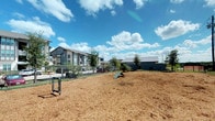 This outdoor dog park is a thoughtfully designed amenity space within a residential community, offering pets and their owners a dedicated area for exercise and recreation. The ground is covered with a generous layer of wood chip mulch, providing a comfortable and low-maintenance surface throughout the open play area. A sturdy black iron perimeter fence encloses the space securely, ensuring the safety of all visitors, while double-gated entry points allow for easy access.

Agility equipment, including a ramp and frame obstacles, is distributed across the park, giving active dogs opportunities for stimulation and play. Conveniently placed waste bag dispensers and disposal stations are positioned throughout, promoting cleanliness and responsible use of the space. Young trees and shrubs dot the landscape, adding a touch of greenery that will mature over time to provide additional shade and visual interest.

The park enjoys open sky views and benefits from abundant natural light. Situated adjacent to a modern residential building with contemporary architectural detailing, the area also offers views of a well-maintained community pool and spa, complete with lounge seating, umbrellas, and a wood-framed pool house entrance. The overall environment is clean, functional, and welcoming for both dogs and their owners.