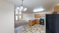 Located at 110-150 39th Ave SE in Minot, North Dakota, this single-floor, one-bedroom, one-bathroom unit offers a practical and comfortable layout. The open-concept kitchen and dining area is well-appointed with warm honey-toned wood cabinetry, a central island with additional storage, stainless steel appliances including a refrigerator and dishwasher, a black range, and a built-in microwave. Stone-patterned tile flooring runs throughout the kitchen and dining space, transitioning seamlessly to plush carpeting in the living room. A multi-bulb chandelier adds a decorative touch above the dining area. The living room is bright and welcoming, featuring multiple windows and a private entry door that allow natural light to fill the space, along with a wall-mounted air conditioning unit and baseboard heating for year-round comfort. The bedroom is a comfortable size, offering a large window with outdoor views, a built-in closet with paneled doors, baseboard heating, and its own wall unit for temperature control. The full bathroom includes a single-sink vanity with wood cabinetry, tile flooring matching the kitchen, a full bathtub with accessibility grab bars, and a toilet. In-unit laundry facilities round out the functional amenities of this well-organized home.
