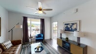 Located at Windridge View in Cincinnati, OH 45243, this single-floor residence offers a thoughtfully designed open-concept layout with contemporary finishes throughout. The living area is bright and welcoming, featuring dark hardwood-style flooring, large windows with dramatic floor-to-ceiling views of the surrounding neighborhood, a ceiling fan with light fixture, and direct access to a private balcony. The space accommodates comfortable seating arrangements anchored by a glass-top coffee table and tufted leather sofa. The kitchen is a standout feature, showcasing rich dark cabinetry, stainless steel appliances including a refrigerator, range, and microwave, a white subway tile backsplash, and a granite-topped island with bar seating beneath brushed nickel pendant lights. A sliding barn door adds a modern-rustic touch near the kitchen and dining area. The bedrooms are carpeted for comfort, each with large windows offering outdoor views and ample natural light. The bathroom is finished with a granite-top dark vanity, a glass-enclosed walk-in shower, and dark hardwood-style flooring, creating a clean and polished aesthetic. Throughout the unit, neutral tones and modern design elements create a cohesive, contemporary ambiance.
