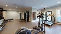 Located at 4300 Jimmy Carter Blvd in Norcross, Georgia, this fitness center offers a dedicated, single-floor exercise room designed to support a well-rounded workout routine. The space is finished with durable speckled rubberized flooring, light gray walls, and elegant crown molding that lends a polished, welcoming atmosphere. Recessed lighting throughout ensures the room is bright and functional at all hours.

Natural light filters in through multiple windows fitted with blinds, allowing users to enjoy outdoor views while exercising. The cardio section is equipped with elliptical trainers, a treadmill, and a stair climber, while the strength training area features a multi-station cable machine, adjustable weight benches, and a dedicated free weight rack stocked with dumbbells and medicine balls. A decorative textured accent wall adds a subtle visual element to the space.

Two dark teal doors provide access to additional areas, and a wall-mounted sanitation station supports cleanliness standards. The overall layout is thoughtfully organized to allow users to move freely between equipment. This fitness center provides a comprehensive and comfortable environment for residents and guests to maintain an active lifestyle.