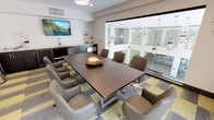 This contemporary conference room is designed to accommodate professional meetings and collaborative gatherings in a polished, well-appointed setting. The centerpiece of the space is a large, dark-wood rectangular boardroom table surrounded by quilted, upholstered chairs on casters, providing both comfort and mobility for attendees.

One wall features a built-in dark cabinetry unit with a sink, countertop workspace, and storage, complemented by a wall-mounted flat-screen television above — making the room well-equipped for presentations and video conferencing. Decorative lanterns and a textured accent wall add visual interest behind the media setup.

Large floor-to-ceiling glass windows and a glass panel wall open the room visually to an interior atrium, flooding the space with abundant natural light and offering views of pendant lighting fixtures and the multi-level building interior. An additional window along another wall frames an outdoor view of palm trees, reinforcing the bright, airy atmosphere.

The flooring consists of patterned carpet tiles in yellow, gray, and brown tones, adding a modern, layered aesthetic. Black-framed glass doors provide a sleek entry point, while decorative mirrors, potted plants, and sculptural accents contribute to a refined yet welcoming ambiance throughout.