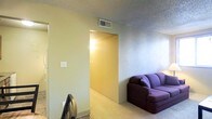 This single-floor space offers a straightforward and functional layout designed for comfortable everyday living. The living area is furnished with a plush sofa and features wall-to-wall carpeting that adds warmth underfoot, while the textured ceiling overhead provides a classic residential character. Neutral-toned walls in soft yellow and light blue hues create a calm, understated atmosphere throughout the space. A central HVAC vent is integrated into the ceiling, ensuring consistent climate control. A wide doorway connects the main living area to an adjoining hallway, which leads further into the interior of the space and is warmly lit by a wall-mounted light fixture. The hallway transition hints at additional rooms beyond, suggesting a practical multi-room floor plan. The open passage between areas promotes an easy flow of movement throughout. Overall, the space presents a clean, no-frills interior that is well-suited for those seeking a comfortable and low-maintenance living environment.