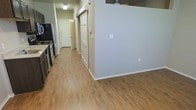 This single-floor residential unit offers a clean, functional layout designed for comfortable everyday living. The interior is unified by warm wood-look luxury vinyl plank flooring that flows seamlessly throughout the open living areas and hallway, contributing to a cohesive and inviting atmosphere.

The kitchen is positioned along one wall and is equipped with standard appliances, including a refrigerator and range, complemented by dark cabinetry that provides ample storage. A partial wall separates the kitchen from the adjoining living area, creating a natural sense of division while maintaining an open feel between the spaces.

A central hallway extends toward the back of the unit, lined with panel doors that lead to individual rooms. The doors feature classic raised-panel detailing, adding a touch of traditional character to the otherwise modern, neutral interior. The walls are finished in a light gray tone that enhances the brightness of the space and serves as a versatile backdrop.

Ceiling-mounted light fixtures provide consistent illumination throughout. The layout is efficient and well-organized, with clear sightlines from the kitchen through to the hallway. Overall, the space presents a tidy, move-in-ready environment with practical finishes suited for residential use.
