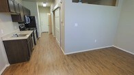 This single-floor residential unit offers a clean, functional layout designed for comfortable everyday living. The interior is unified by warm wood-look luxury vinyl plank flooring that flows seamlessly throughout the open living areas and hallway, contributing to a cohesive and inviting atmosphere.

The kitchen is positioned along one wall and is equipped with standard appliances, including a refrigerator and range, complemented by dark cabinetry that provides ample storage. A partial wall separates the kitchen from the adjoining living area, creating a natural sense of division while maintaining an open feel between the spaces.

A central hallway extends toward the back of the unit, lined with panel doors that lead to individual rooms. The doors feature classic raised-panel detailing, adding a touch of traditional character to the otherwise modern, neutral interior. The walls are finished in a light gray tone that enhances the brightness of the space and serves as a versatile backdrop.

Ceiling-mounted light fixtures provide consistent illumination throughout. The layout is efficient and well-organized, with clear sightlines from the kitchen through to the hallway. Overall, the space presents a tidy, move-in-ready environment with practical finishes suited for residential use.