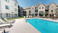 Located at 2701 Fletcher Ave in Lincoln, Nebraska, this outdoor pool area offers a well-appointed recreational retreat set within a modern residential community. The centerpiece is a generously sized swimming pool with crystal-clear blue water, entry steps, and stainless steel handrails for easy access. The expansive concrete pool deck surrounds the water on multiple sides, providing ample space for lounging and movement.

A row of white reclining sun loungers lines the poolside, offering residents comfortable spots to relax and soak up the Nebraska sun. Teal-accented patio umbrellas provide shaded seating areas throughout the deck, while a covered brick gazebo with an open-air dining table and chairs serves as a gathering space for outdoor meals and socializing.

The pool area is fully enclosed by a black wrought-iron security fence, ensuring privacy and safety. Lush green lawn areas are visible along the perimeter, adding a touch of natural landscaping to the hardscape setting. The surrounding multi-story residential buildings feature board-and-batten siding in neutral tones, pitched rooflines, and private balconies that overlook the pool courtyard, creating an inviting, community-centered atmosphere beneath open Nebraska skies.
