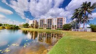 This multi-story residential building is set within a well-maintained landscape that immediately captures attention. The exterior showcases a classic Florida architectural style, featuring a warm, neutral facade with rows of private balconies on each floor, offering residents elevated views of the surrounding environment. The building rises several stories, providing a substantial and commanding presence on the property. Lush, manicured lawns extend outward from the base of the structure, transitioning to the banks of a tranquil pond that reflects the building and the dramatic, cloud-filled sky above. Mature palm trees are scattered throughout the grounds, reinforcing the tropical character of the setting and providing natural shade and visual texture. The pond adds a peaceful, scenic element to the outdoor environment, enhancing the overall ambiance of the property. The grounds are well-kept, with trimmed hedgerows and green open spaces that contribute to a park-like atmosphere. The combination of the waterfront setting, tropical landscaping, and multi-level balconies creates a residential environment that is both visually appealing and conducive to outdoor enjoyment.