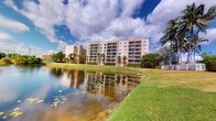 This multi-story residential building is set within a well-maintained landscape that immediately captures attention. The exterior showcases a classic Florida architectural style, featuring a warm, neutral facade with rows of private balconies on each floor, offering residents elevated views of the surrounding environment. The building rises several stories, providing a substantial and commanding presence on the property. Lush, manicured lawns extend outward from the base of the structure, transitioning to the banks of a tranquil pond that reflects the building and the dramatic, cloud-filled sky above. Mature palm trees are scattered throughout the grounds, reinforcing the tropical character of the setting and providing natural shade and visual texture. The pond adds a peaceful, scenic element to the outdoor environment, enhancing the overall ambiance of the property. The grounds are well-kept, with trimmed hedgerows and green open spaces that contribute to a park-like atmosphere. The combination of the waterfront setting, tropical landscaping, and multi-level balconies creates a residential environment that is both visually appealing and conducive to outdoor enjoyment.