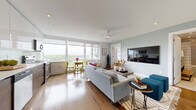 This contemporary single-floor residence is thoughtfully designed with a clean, modern aesthetic and warm natural finishes. The open-plan living and dining area is the heart of the home, anchored by wide-plank hardwood floors and floor-to-ceiling windows that flood the space with natural light and offer sweeping elevated views of a lush green canopy. A ceiling fan, recessed lighting, and soft sage-toned walls create a relaxed yet refined ambiance. The living area flows effortlessly into a compact dining nook and connects to a sleek, galley-style kitchen outfitted with rich wood cabinetry, high-gloss white upper cabinets, a white quartz countertop, a glass tile backsplash, and stainless steel appliances including a dishwasher and built-in microwave. The bedroom is a bold and cozy retreat, featuring a striking magenta accent wall, hardwood floors, a large window with treetop views, and a well-organized mirrored closet with built-in shelving and drawers. The bathroom is finished in neutral stone-look tile with a deep soaking tub, a multi-jet shower system, a floating wood-toned vanity, and a wooden slatted bench — offering a serene, spa-like experience. In-unit laundry is also present, adding everyday convenience to this well-appointed residence.