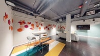 Located at 1225 Santa Fe Dr in Denver's creative Santa Fe Arts District, this community room is a thoughtfully designed single-floor gathering space offering a compelling blend of industrial aesthetics and colorful modern design. The open layout accommodates multiple functional zones, creating a versatile environment suited for social events, meetings, and casual gatherings. Exposed ductwork and track lighting traverse the dark ceiling, reinforcing an urban, loft-inspired character throughout the space. The lounge area features contemporary gray sofas and a plush area rug, providing a comfortable setting for relaxed conversation. A high-top bistro table with wire-frame bar stools occupies a dedicated nook, anchored by a vibrant multi-colored tile carpet in bold stripes of teal, orange, and yellow. Walls throughout are adorned with a striking geometric hexagonal mural in warm reds, oranges, and magentas, adding a dynamic artistic focal point. The fully equipped kitchen includes sleek dark gray cabinetry, a subway tile backsplash, white quartz countertops, stainless steel appliances, and a peninsula with bar seating — ideal for catering or communal use. Large-format dark tile flooring unifies the space, while structural columns add architectural character. Track and pendant lighting ensure the room is well-illuminated throughout.