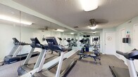 Located at 6741 C Avenue NE in Cedar Rapids, Iowa, this fitness center offers a practical and fully equipped exercise space designed to support a well-rounded workout routine. The single-room facility is finished with light sage green walls, a textured ceiling, and durable carpet flooring, creating a clean and comfortable environment for physical activity. Ceiling fans provide air circulation throughout the space, complementing the overhead lighting. A defining architectural feature is the large wall-to-wall mirror installation along one side of the room, which visually expands the space and enhances functionality during workouts. The room is equipped with multiple treadmills, an elliptical machine, and a stationary bike, providing ample cardio options. Strength training is well represented with a weight bench, a rack of dumbbells in varying sizes, and medicine ball storage. A wall-mounted television is positioned near the entry for entertainment during exercise sessions. The entry features a glass-paneled double door with exterior views, allowing natural light to filter into the space. Additional amenities include a hand sanitizer station, sanitation supplies for equipment cleaning, and a fire extinguisher, reflecting attention to safety and hygiene. Interior doors provide access to additional utility or storage areas.