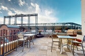 The Battery Rooftop offers a remarkable outdoor space that combines urban sophistication with recreational comfort. This single-floor terrace boasts an impressive layout with multiple seating arrangements, including dining tables, lounge chairs, and casual seating options. The flooring consists of a stylish mix of wooden decking in various patterns, creating visual interest and defining different areas.

A standout feature is the breathtaking view of a nearby stadium, its impressive steel framework dominating the skyline. This proximity to a major sports venue adds a unique ambiance to the space. The cityscape beyond provides a stunning backdrop, with modern skyscrapers visible in the distance.

The rooftop is equipped with practical amenities, including a built-in grill for outdoor cooking and entertaining. Safety features are evident, with sturdy railings along the perimeter and clear signage indicating rules such as no smoking. Planters with greenery add a touch of nature to the urban setting.

For entertainment and information, a large outdoor screen is mounted on the wall, possibly for viewing events or presentations. The space appears to be designed for versatility, suitable for both casual relaxation and organized events.

Weather protection seems to be considered, with some areas potentially covered or shaded, though not visible in the provided images. The open layout allows for ample natural light and air circulation, making it an ideal spot for enjoying fair weather days high above the city streets.