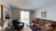 Portage Point is a single-floor residential space designed for comfortable, functional living. The living room serves as the heart of the unit, offering a welcoming and well-appointed layout. A large sliding glass door with vertical blinds fills the room with natural light while providing direct access to an outdoor balcony, creating a seamless connection between the interior and exterior. A wall-mounted air conditioning unit ensures year-round climate comfort. The living area features neutral-toned walls and textured ceilings that lend the space a classic, understated character. Plush carpeting underfoot adds warmth to the room, complementing the cozy seating arrangement that includes a leather recliner and a generous upholstered sofa accented with decorative pillows. A dark wood coffee table anchors the space, adding contrast to the light surroundings. Framed wall art contributes a personalized touch to the overall ambiance. The space strikes a balance between practicality and comfort, making it well-suited for relaxed everyday living. Its single-floor layout offers straightforward accessibility throughout, while the outdoor balcony provides an added layer of openness and connection to the surrounding environment at Portage Point.