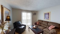 Portage Point is a single-floor residential space designed for comfortable, functional living. The living room serves as the heart of the unit, offering a welcoming and well-appointed layout. A large sliding glass door with vertical blinds fills the room with natural light while providing direct access to an outdoor balcony, creating a seamless connection between the interior and exterior. A wall-mounted air conditioning unit ensures year-round climate comfort. The living area features neutral-toned walls and textured ceilings that lend the space a classic, understated character. Plush carpeting underfoot adds warmth to the room, complementing the cozy seating arrangement that includes a leather recliner and a generous upholstered sofa accented with decorative pillows. A dark wood coffee table anchors the space, adding contrast to the light surroundings. Framed wall art contributes a personalized touch to the overall ambiance. The space strikes a balance between practicality and comfort, making it well-suited for relaxed everyday living. Its single-floor layout offers straightforward accessibility throughout, while the outdoor balcony provides an added layer of openness and connection to the surrounding environment at Portage Point.
