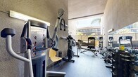 Located at 1348 N Sierra Bonita Ave in the heart of Los Angeles, this fitness center offers a dedicated exercise room thoughtfully equipped for a comprehensive workout experience. The space features notably high ceilings that contribute to an open, airy atmosphere, with textured stucco walls characteristic of the building's architectural style. A large open-air section at one end of the room invites natural light and outdoor views of swaying palm trees, creating a bright and energizing environment that blends indoor and outdoor fitness. The rubber-floored exercise room is well-stocked with a wide variety of equipment to support both cardiovascular and strength training. Cardio offerings include treadmills, an elliptical trainer, and stationary bikes. For strength training, the room provides a multi-station cable machine, a leg press, a weight bench, a squat rack, and an extensive dumbbell rack ranging across multiple weight increments, supplemented by kettlebells. A wall-mounted mirror and flat-screen TV enhance the workout environment. Fluorescent lighting ensures the interior is well-lit throughout. The space is efficiently organized to maximize usability within its footprint, making it a practical and fully functional fitness facility.