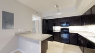 This single-floor residence showcases a thoughtfully designed interior with a distinctly modern aesthetic. The kitchen is a standout feature, outfitted with bold, floor-to-ceiling dark cabinetry that creates a striking contrast against the light-toned walls and tile flooring. Granite countertops with a subtle speckled pattern add texture and elegance, while the full suite of black appliances — including a refrigerator with ice dispenser, electric range, built-in microwave, and dishwasher — delivers a cohesive, contemporary look. A peninsula counter extends into the space, offering additional prep area and casual seating potential, making it an inviting hub for both cooking and entertaining. Track lighting mounted on the ceiling provides focused illumination throughout the kitchen. Light-colored tile flooring runs throughout, enhancing the sense of openness and brightness. A glimpse into an adjacent area reveals a laundry space, adding to the unit's functional appeal. The overall ambiance is clean, polished, and modern, with a well-considered balance of dark and light tones that gives the space a refined, upscale character.